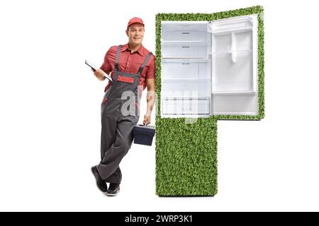 Servicemitarbeiter mit Klemmbrett und Werkzeugkasten, der sich gegen einen grünen, nachhaltigen Kühlschrank lehnt, isoliert auf weißem Hintergrund Stockfoto