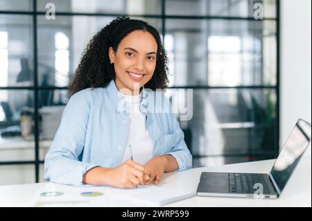 Positive hispanische oder brasilianische Frau mit lockigem Haar in einem Hemd, Manager, Sekretärin, sitzt am Arbeitsplatz im Büro vor einem Laptop, macht Notizen in einem Notebook, sieht aus und lächelt in die Kamera Stockfoto