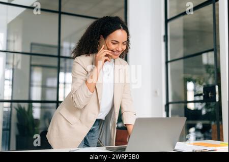 Telefonieren. Geschäftige, hübsche brasilianische oder hispanische Geschäftsfrau, Sekretärin, Managerin, steht in einem modernen Büro neben einem Schreibtisch, arbeitet in einem Laptop und führt ein Smartphone-Gespräch, lächelt Stockfoto