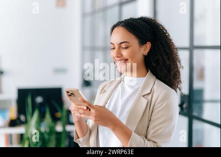 Eine schöne positive brasilianische oder hispanische junge Frau mit lockigen Haaren in einer eleganten Jacke, steht in einem modernen Büro, benutzt ihr Smartphone, sendet Nachrichten mit Freunden in sozialen Netzwerken, lächelt Stockfoto