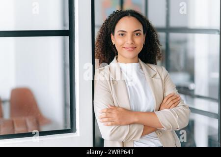 Porträt einer selbstbewussten, hinreißenden, erfolgreichen hispanischen oder brasilianischen jungen Frau mit lockigem Haar, steht in einem modernen Büro mit überkreuzten Armen, blickt in die Kamera und lächelt freundlich Stockfoto
