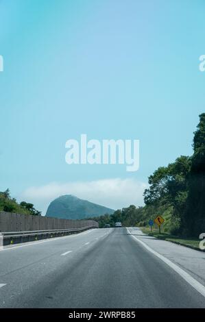 Blick auf KM 5 der Via Lagos Highway mit einem großen Berg, der von dichter Atlantikwaldvegetation und Wolken im Hintergrund unter dem klaren blauen Sommerhimmel bedeckt ist. Stockfoto