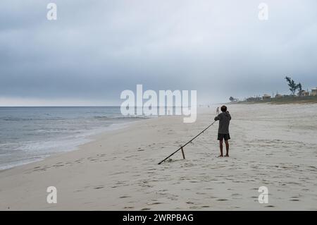 Ein Fischer, der seine Fischrute am weißen Sand des Praia Seca Strandes vorbereitet, in der Nähe des blauen Wassers des Atlantiks unter dem Wolkenhimmel am Sommernachmittag. Stockfoto