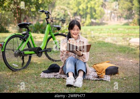 Eine positive, glückliche junge Asiatin liest ein Buch und schlürft Kaffee, während sie sich in einem grünen Park entspannt. Menschen- und Lifestyle-Konzepte Stockfoto