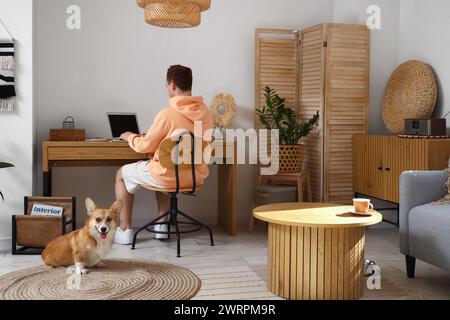 Rothaariger junger Mann mit Laptop und süßem Corgi-Hund, der zu Hause arbeitet Stockfoto