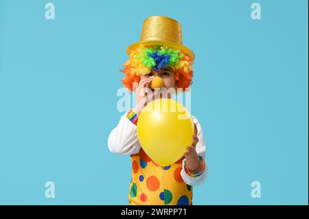 Lustiger kleiner Junge im Clown-Kostüm mit Hut, der Ballon auf blauem Hintergrund hält. Feierlichkeiten zum Aprilfeiertag Stockfoto