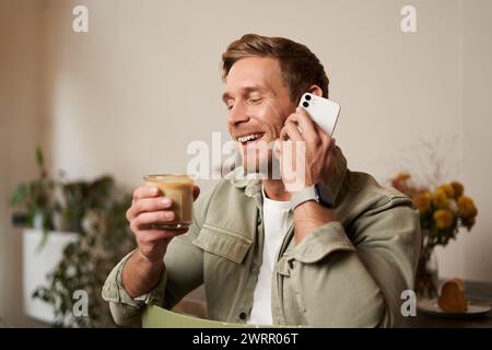 Porträt eines jungen gutaussehenden Mannes, der am Telefon spricht, Kaffee trinkt und lacht. Ein Kerl sitzt im Café und hat eine nette, angenehme Unterhaltung bei einer Tasse Stockfoto