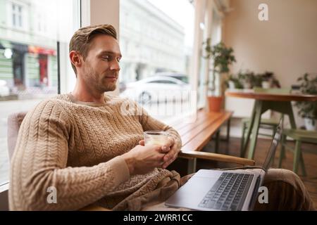 Porträt eines männlichen Unternehmers, digitaler Nomade, der mit einem Glas Cappuccino im Café sitzt, einen Laptop anschaut, Videos ansieht, Online-Meetings teilnimmt Stockfoto