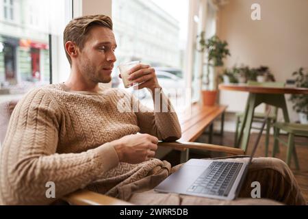 Working People und Public Space Konzept. Junger gutaussehender Mann, ux ui Designer sitzt mit Laptop im Café, trinkt Cappuccino und schaut mit auf den Bildschirm Stockfoto