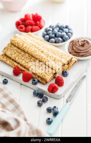 Frische Crepes mit frischen Beeren serviert auf einem weißen Gericht neben Himbeeren, Heidelbeeren und Schokoladenaufstrich. Stockfoto