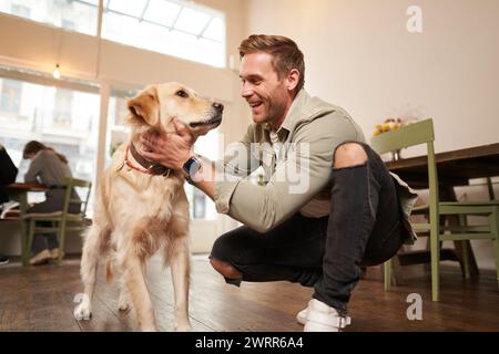 Porträt eines glücklichen jungen Mannes, der mit seinem goldenen Retriever ins Café geht. Hübscher Kerl, der Hund in einem haustierfreundlichen Café streichelt Stockfoto