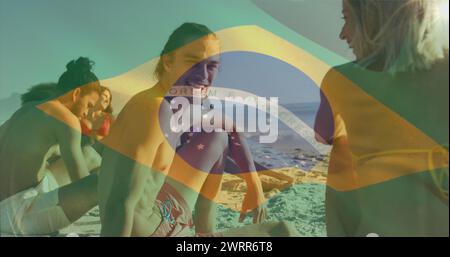 Bild der Flagge brasiliens, die über verschiedene Freunde schwenkt, während sie am Strand sitzen Stockfoto