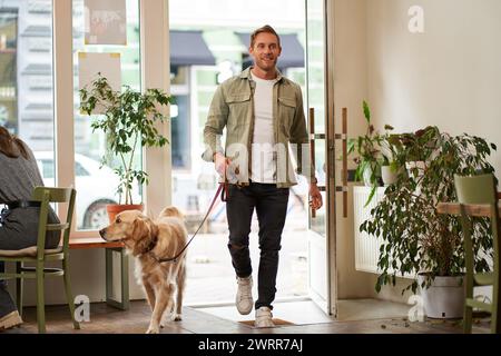 Porträt eines gutaussehenden jungen Mannes, der mit seinem Hund an der Leine ins Café geht, das haustierfreundliche Café betritt und die Tür öffnet Stockfoto