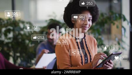 Bild von Benachrichtigungen in sozialen Medien über glückliche birassische Frau, die ein Tablet im Büro verwendet Stockfoto