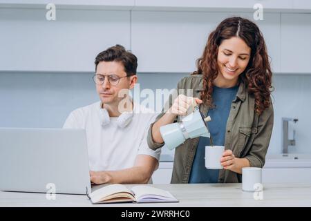 Eine Frau gießt Kaffee, während ein Mann an einem Laptop arbeitet, und beide genießen eine entspannte Arbeit von zu Hause aus. Ihre bequeme Einstellung fördert Produktivität und Balance. Kaffeepause Während Der Arbeit Von Zu Hause Aus Stockfoto