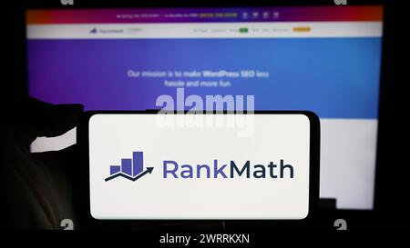 Person mit Handy mit Logo der indischen WordPress SEO-Firma Rank Math vor der Business-Webseite. Schwerpunkt auf dem Display des Telefons. Stockfoto