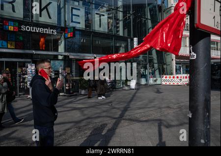 05.03.2024, Berlin, Deutschland, Europa - Straßenszene zeigt einen Mann mit Zigarette und rotem Handy neben einer roten Plastikfolie, die in den Wind bläst. Stockfoto