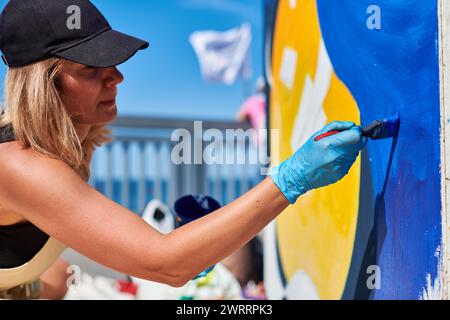 Erwachsene Malerin in schwarzer Kappe malt leidenschaftlich Bild mit Pinsel für die Straßenausstellung im Freien mit leuchtenden Farben, visuelles Spektakel durch Stockfoto