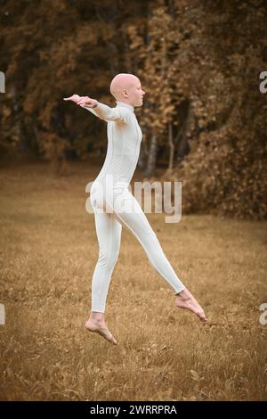 Das Porträt der jungen, haarlosen Mädchen-Ballerina mit Alopezie im engen weißen Anzug bereitet sich darauf vor, auf den Herbstrasen im Park zu springen und symbolisiert die Überwindung Stockfoto