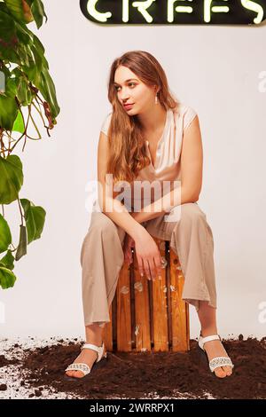 Eine junge, blonde Frau mit langen Haaren trägt ein sommerliches Outfit. Sie sitzt auf einer Weinkiste, die auf dem Boden steht. Zu ihrer Linken ist eine kletternde pla Stockfoto