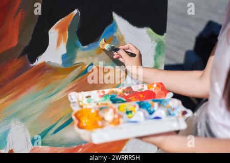 Weibliche Malerhand malt leidenschaftlich mit Pinsel für die Straßenausstellung im Freien mit leuchtenden Farben, Nahaufnahme Stockfoto