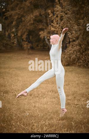 Das Porträt der jungen, haarlosen Mädchen-Ballerina mit Alopezie im engen weißen Anzug bereitet sich darauf vor, auf den Herbstrasen im Park zu springen und symbolisiert die Überwindung Stockfoto