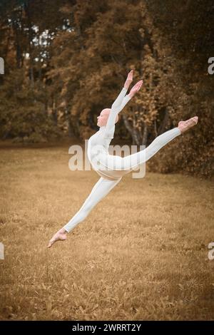 Porträt der jungen, haarlosen Mädchen Ballerina mit Alopezie im engen weißen Anzug springt auf den Herbstrasen im Park und symbolisiert die Bewältigung der Herausforderungen Stockfoto