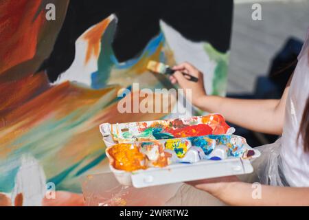 Weibliche Malerhand malt leidenschaftlich mit Pinsel für die Straßenausstellung im Freien mit leuchtenden Farben, Nahaufnahme Stockfoto