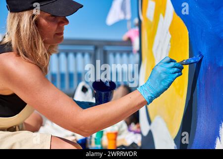 Erwachsene Malerin in schwarzer Kappe malt leidenschaftlich Bild mit Pinsel für die Straßenausstellung im Freien mit leuchtenden Farben, visuelles Spektakel durch Stockfoto