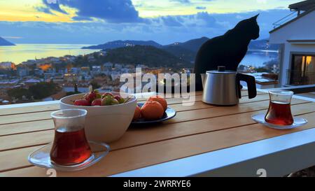 Romantische Teeparty bei Sonnenuntergang für Paare. Frische Erdbeeren und Mandarinen, schwarzer Tee in türkischer Tasse. Niemand, schwarze Katze auf der Terrasse mit der Stadt am Meer Stockfoto