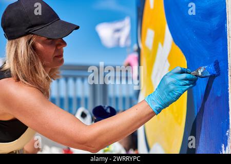 Erwachsene Malerin in schwarzer Kappe malt leidenschaftlich Bild mit Pinsel für die Straßenausstellung im Freien mit leuchtenden Farben, visuelles Spektakel durch Stockfoto