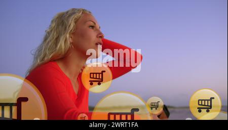 Bild von Einkaufsymbolen über der Mitte einer kaukasierin, die am Strand ein Smartphone benutzt Stockfoto