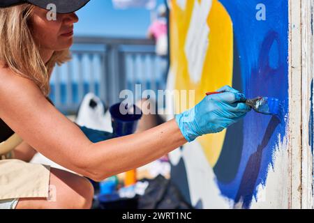 Erwachsene Malerin in schwarzer Kappe malt leidenschaftlich Bild mit Pinsel für die Straßenausstellung im Freien mit leuchtenden Farben, visuelles Spektakel durch Stockfoto