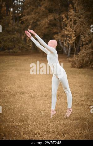 Das Porträt der jungen, haarlosen Mädchen-Ballerina mit Alopezie im engen weißen Anzug bereitet sich darauf vor, auf den Herbstrasen im Park zu springen und symbolisiert die Überwindung Stockfoto