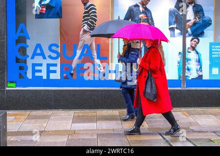 Frauen mit langem Mantel und rotem Regenschirm in Preston, Lancashire. Wetter in Großbritannien. März 2024. Regnerischer Start in den Tag für Shopper im Stadtzentrum. Wind mit heftigeren Regenfällen. Credit; MediaWorldImages/AlamyLiveNews Stockfoto