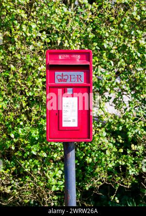 Ein roter Briefkasten auf einem Pol an einem Grasrand vor einer Hecke, in einem ländlichen Dorf in Lancashire Stockfoto