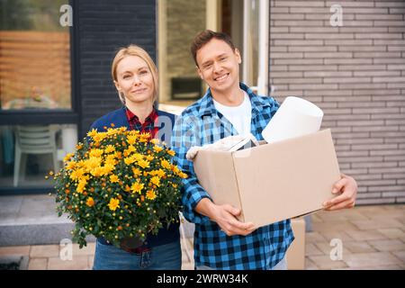 Hüpfen Sie das Bild von einem freudigen Mann mit Pappkarton und einer lächelnden Frau, die Blumen hält, wenn sie beim Umzug in der Nähe des Gebäudes steht Stockfoto