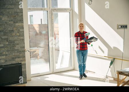 Foto in voller Länge einer fröhlichen Frau, die in einem unvollendeten Raum steht, während sie Maltablett und Walze in den Händen hält Stockfoto