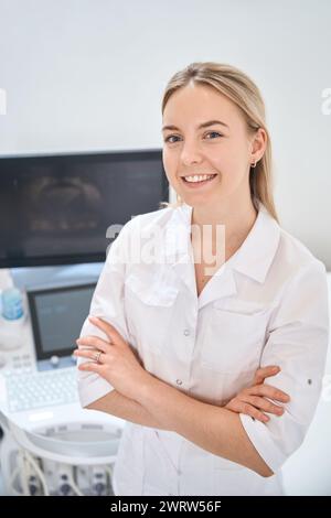 Ziemlich hoch qualifizierte Ultraschallfachfrau lächelnd in der Nähe des Ultraschallgeräts, Klinik schlägt alle Arten von Ultraschall für Menschen vor, Gesundheitsauto Stockfoto