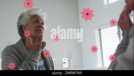 Bild von rosa Blumen über glücklicher birassischer Seniorin, die zu Hause im Spiegel Make-up aufsetzt Stockfoto