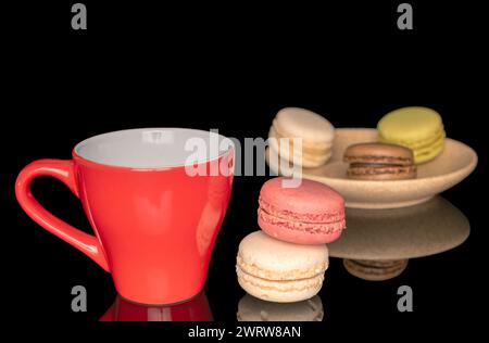 Mehrere süße Makronen auf einer Keramikuntertasse mit einer roten Tasse, Makro, isoliert auf schwarzem Hintergrund. Stockfoto