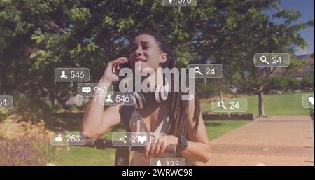 Bild von Mediensymbolen über lächelnder birassischer Frau mit Roller, die auf dem Smartphone im Park spricht Stockfoto