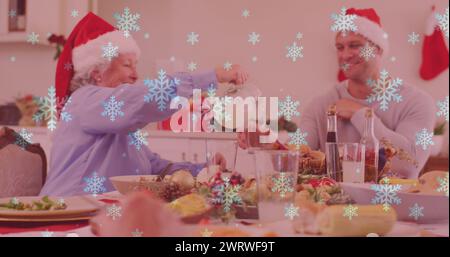 Bild von Schnee über lächelnde kaukasische Familie mit weihnachtsmützen beim Abendessen Stockfoto