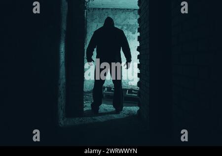 Horrorfoto eines Mannes in schwarzer Kapuze, Stalker oder Mörder, der in einem dunklen verlassenen Gebäude steht. Stockfoto