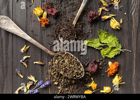 Zusammensetzung von trockenem schwarzem und grünem Tee in klassischen Löffeln und trockenem Hibiskus, Kalendula, Lavendelblumen auf dunklem Holzhintergrund, Nahaufnahme von oben. Stockfoto
