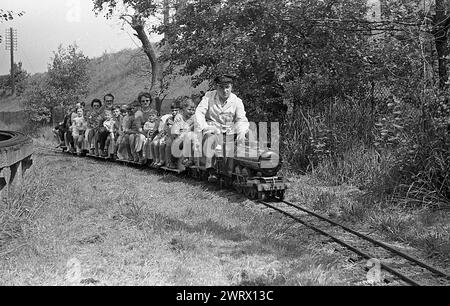1970er Jahre, historisch, Erwachsene und Kinder fahren auf einer Miniatureisenbahn, England, Großbritannien, Zugführer im Overall und Hut, der die kleine Dampfeisenbahn auf der Außenbahn fährt. Stockfoto