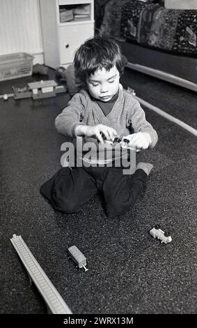 1970er Jahre, historisch, ein kleiner Junge, der in seinem Schlafzimmer sitzt und mit seinem hölzernen Spielzeugzug spielt. Stockfoto