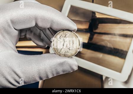 Foto einer männlichen Hand in weißen Handschuhen, die eine sowjetische Silbermünze 1924 Halb Rubel auf einem Stapel Bücher und einem Lupenhintergrund hält. Numismat Stockfoto