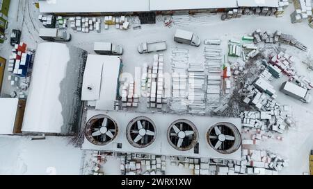 Drohnenfotografie von Kühlventilatoren im industriellen Maßstab und Baumaterial in einem Freiluftlager, das während des bewölkten Winters von Schnee bedeckt ist Stockfoto