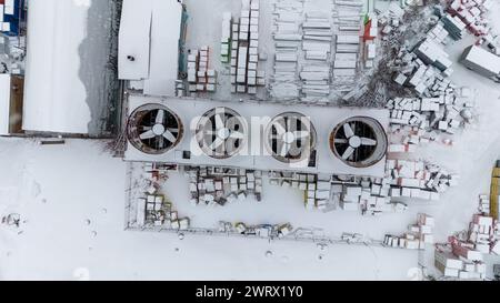 Drohnenfotografie von Kühlventilatoren im industriellen Maßstab und Baumaterial in einem Freiluftlager, das während des bewölkten Winters von Schnee bedeckt ist Stockfoto